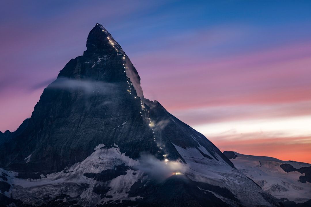 Das Matterhorn in der Abenddämmerung, beleuchtet von einer Lichterkette, die vor einem farbenfrohen Himmel den Hang hinaufführt, erinnert an den Charme der historischen Landschaften Aachens.