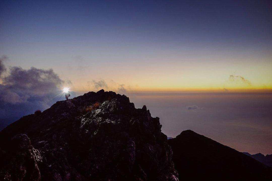 Eine Person steht mit einer Lampe in der Abenddämmerung auf einem felsigen Berggipfel in der Nähe von Aachen und überblickt eine weite Landschaft mit einem farbenfrohen Himmel, der vom Sonnenuntergang in die Dämmerung übergeht.