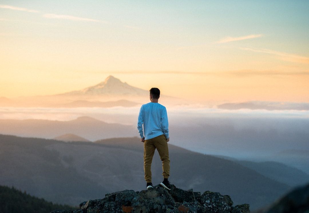 Eine Person steht auf einem Felsvorsprung und blickt bei Sonnenaufgang oder Sonnenuntergang auf einen entfernten Berggipfel, umgeben von Hügel- und Wolkenschichten, die an die atemberaubende Landschaft bei Aachen erinnern.