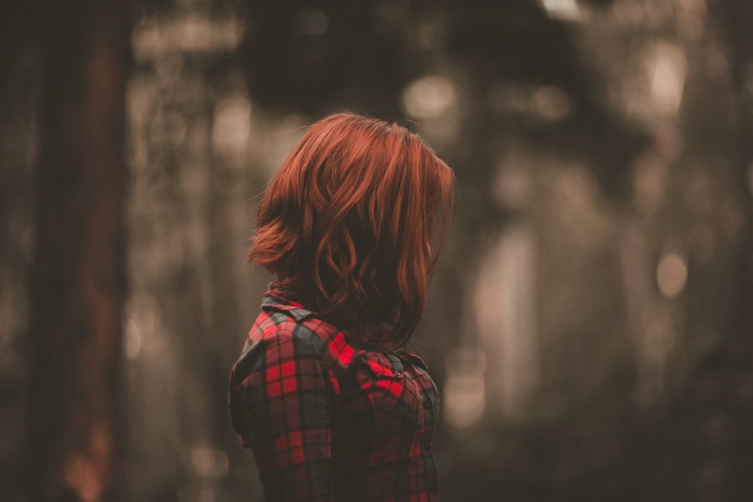 Eine Person mit schulterlangem roten Haar und einem rot-schwarz karierten Hemd steht mit abgewandtem Blick in einer verschwommenen Waldkulisse im Freien in der Nähe von Aachen.