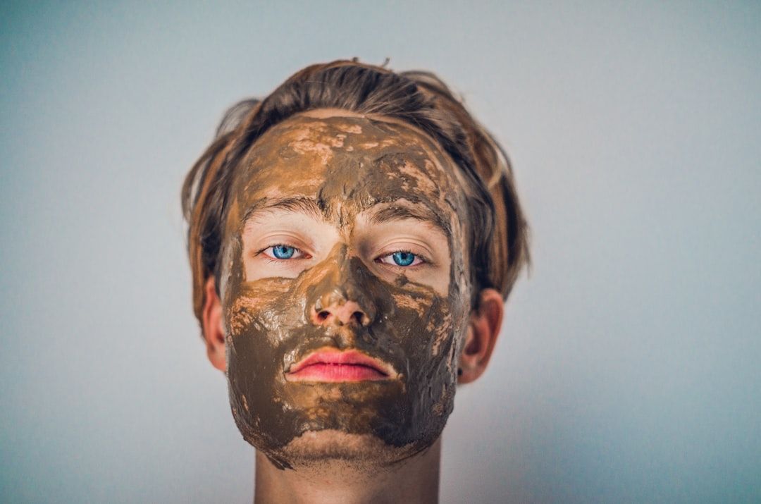 Eine Person mit blauen Augen blickt nach vorne. Sie trägt eine braune Gesichtsmaske. Ihre Haare sind leicht zerzaust und der schlichte Hintergrund lässt auf einen ruhigen Tag in Aachen schließen.