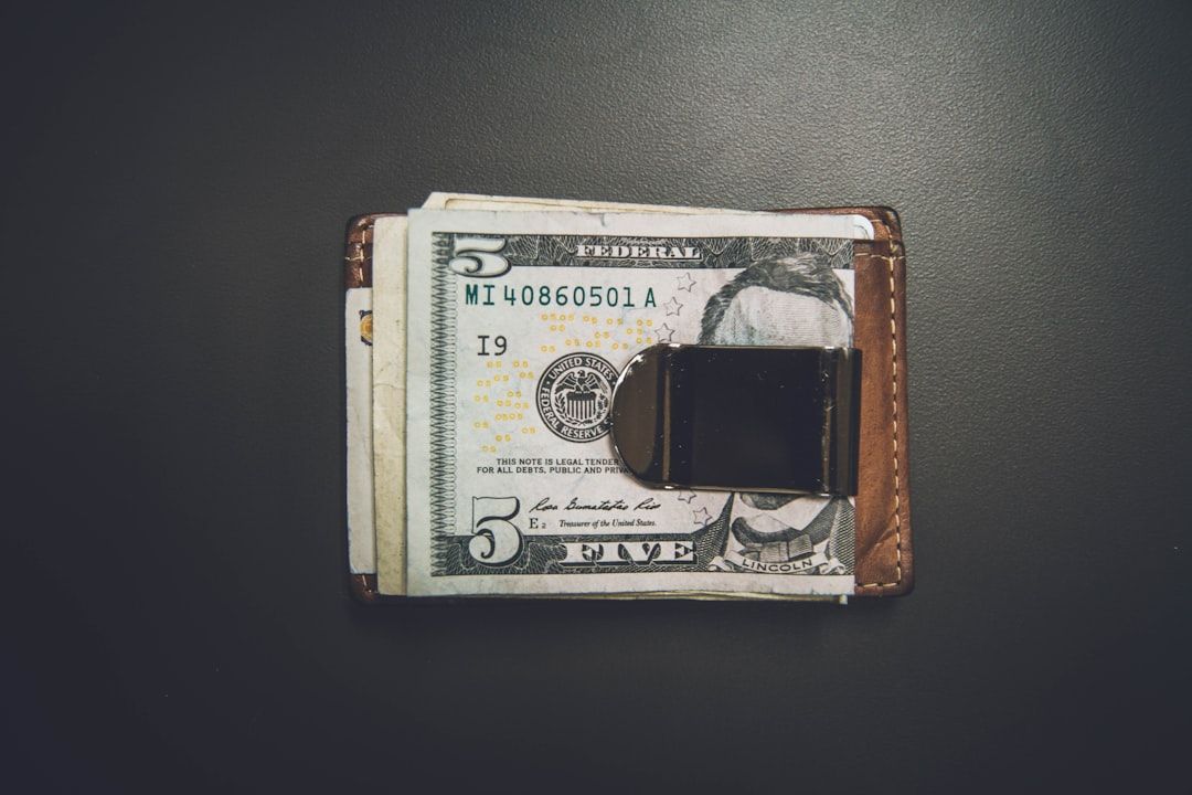 Eine Brieftasche mit Metallclip, in der sich mehrere Fünf-Dollar-Scheine befinden, liegt auf einer dunklen Oberfläche und strahlt die zeitlose Eleganz der Aachener Handwerkskunst aus.