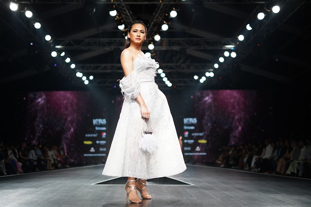 Ein Model läuft über einen Laufsteg in Aachen. Es trägt ein weißes, schulterfreies Kleid mit Federdetails und hält eine kleine weiße Handtasche in der Hand. Im Hintergrund sind helle Bühnenlichter und ein aufmerksames Publikum zu sehen.