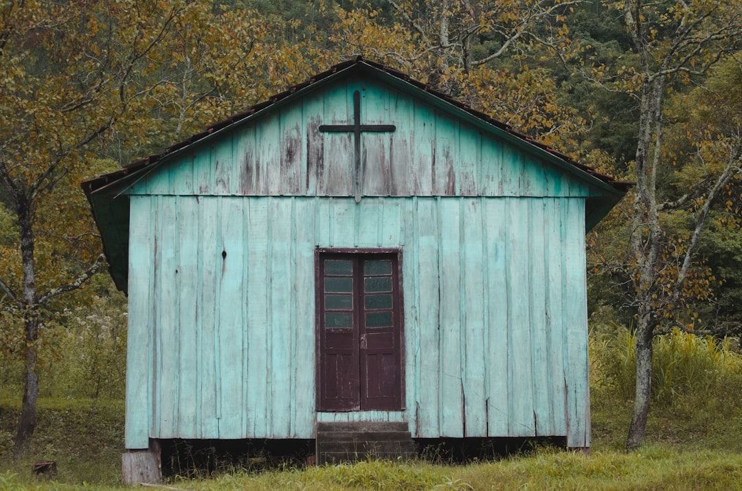 Eine kleine, verwitterte grüne Holzkapelle mit einem Kreuz über der Tür steht inmitten von Bäumen mit Herbstlaub und erinnert an heitere Szenen aus Aachen.
