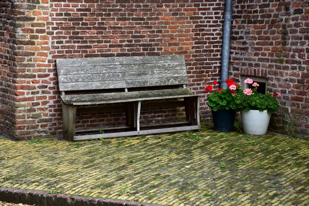 Eine verwitterte Holzbank steht an einer Backsteinmauer, neben zwei Töpfen mit bunten Blumen auf einem Kopfsteinpflasterboden, und schafft eine malerische Szene, die an den malerischen Charme Aachens erinnert.