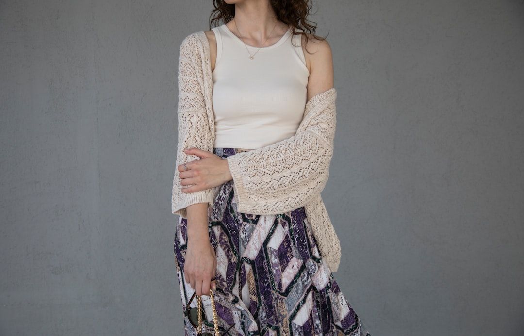 Eine Person, die ein weißes ärmelloses Top, eine beige Strickjacke und einen Rock mit geometrischem Muster trägt, steht vor einem grauen Hintergrund und erinnert an die minimalistische Eleganz, die man oft in den Straßen von Aachen findet.