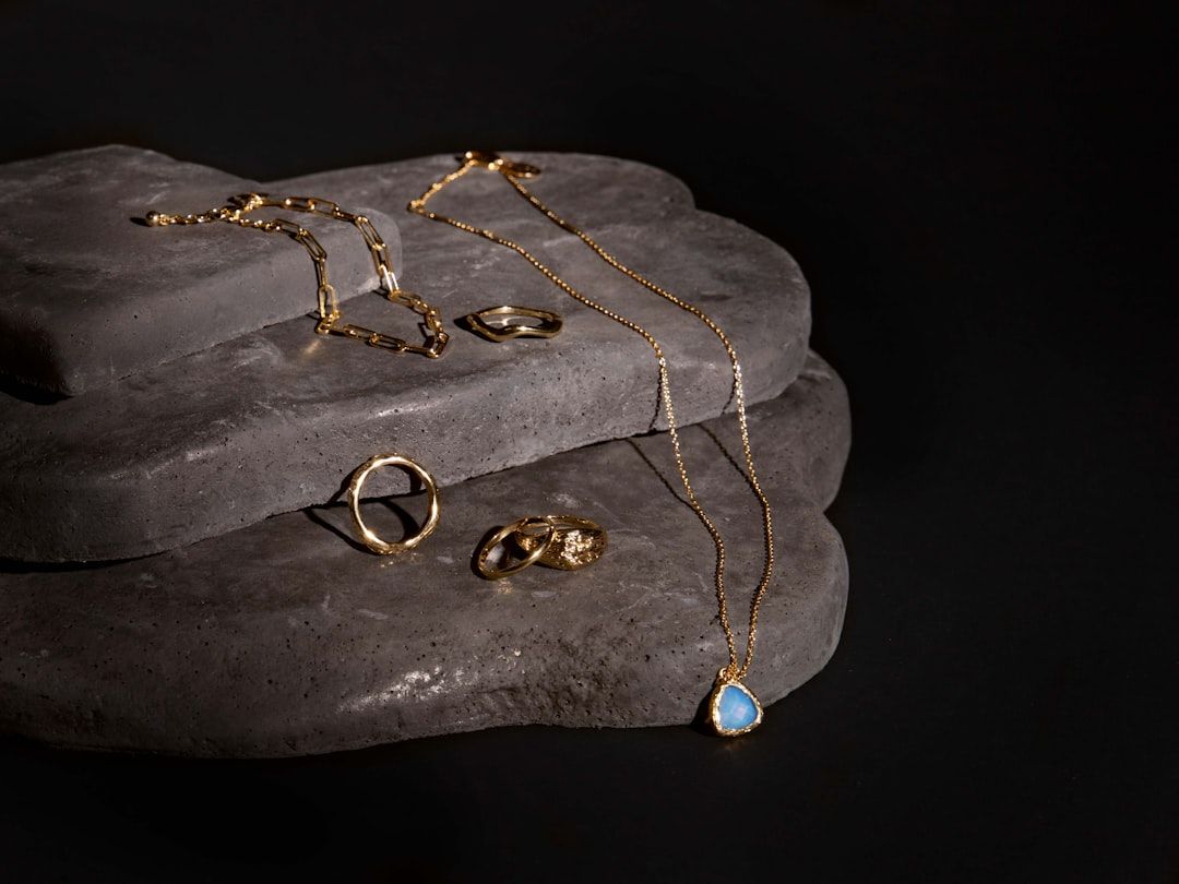 Eine Sammlung von Goldschmuck, darunter eine Halskette mit blauem Anhänger, Armbänder und Ringe, inspiriert vom Reichtum Aachens, präsentiert auf gestapelten Schieferplattformen vor dunklem Hintergrund.