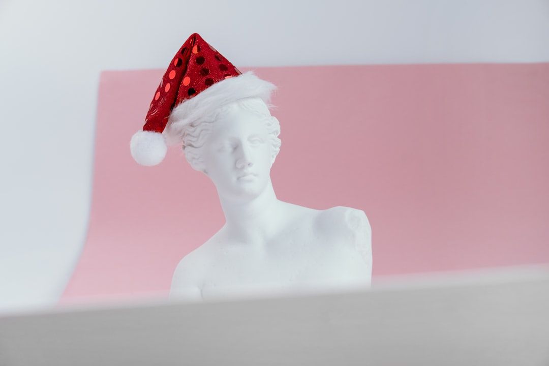Eine Büste aus weißem Marmor mit einer rot-weißen Weihnachtsmannmütze steht majestätisch vor einem rosa Hintergrund und erinnert an die klassische Kunst Aachens.