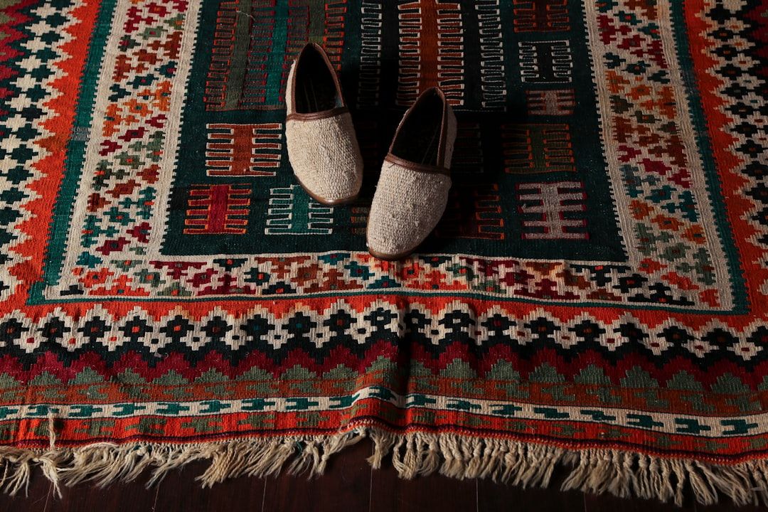 Gemusterter Textilteppich mit Fransen auf dem Boden, dessen geometrische Muster in Rot, Grün und Weiß an die reiche Geschichte Aachens erinnern. In der Mitte des Teppichs steht ein Paar Slipper aus cremefarbenem Canvas mit dunkler Verzierung.