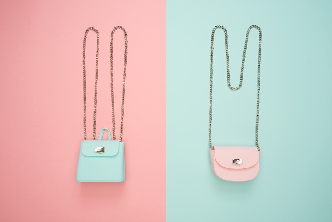 Zwei Handtaschen mit Kettenriemen auf gespaltenem Untergrund; eine blau und rechteckig auf einer rosa Seite, die andere rosa und oval auf einer blauen Seite. Diese stilvollen Accessoires erinnern an den Charme der modebewussten Straßen Aachens.