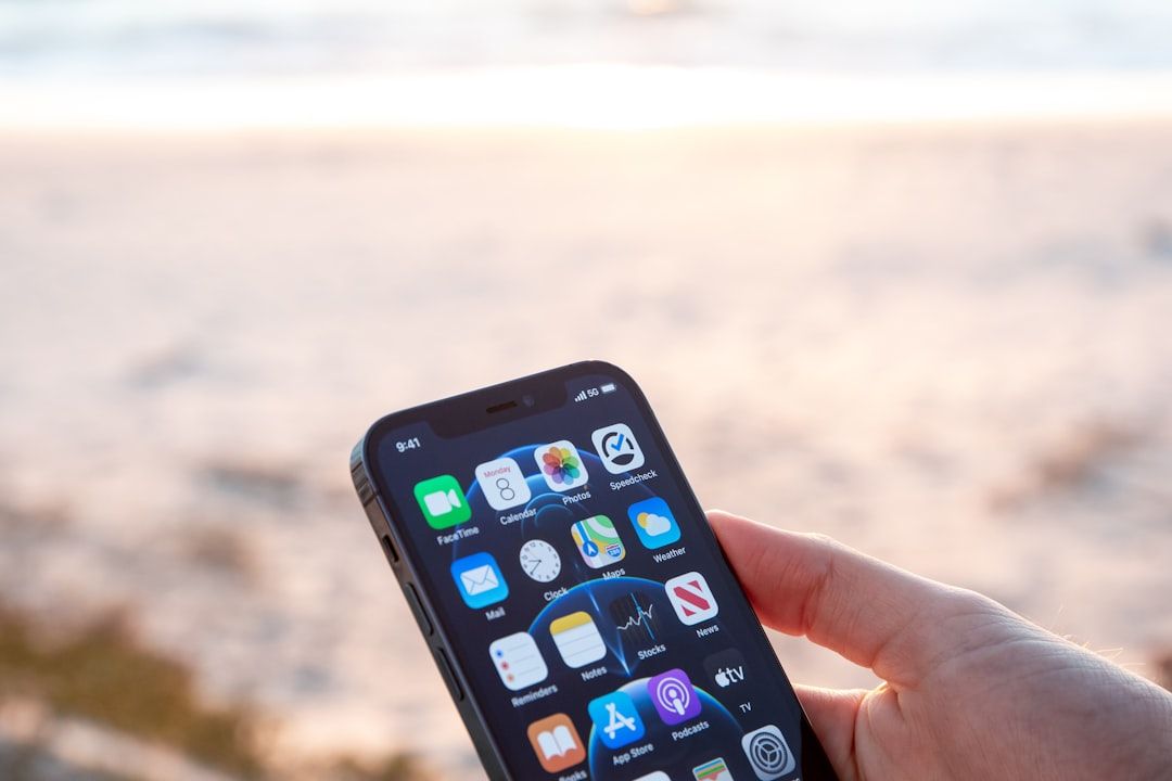 Person, die ein Smartphone mit verschiedenen App-Symbolen auf dem Bildschirm vor einem verschwommenen Strandhintergrund hält und vielleicht an ihre letzte Reise nach Aachen zurückdenkt.