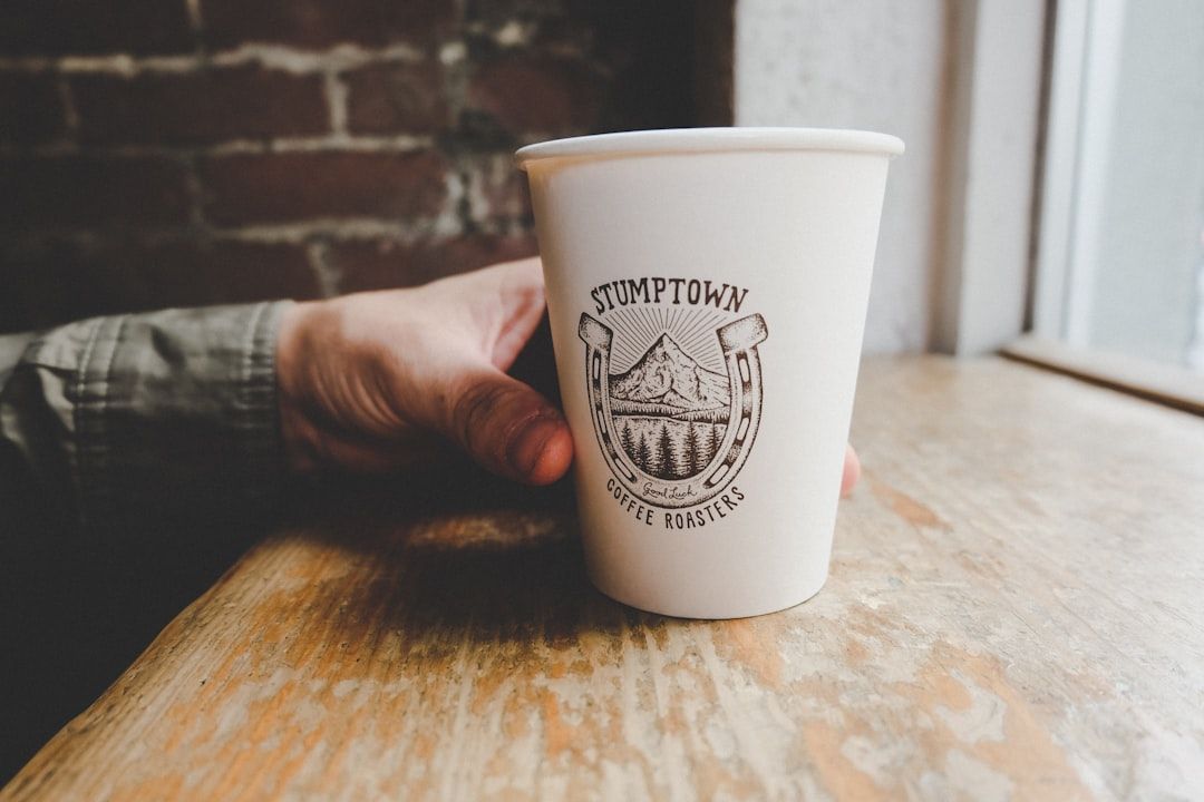 Eine Hand, die einen weißen Kaffeebecher aus Pappe mit dem Logo von „Stumptown Coffee Roasters“ hält und auf einer Holzoberfläche neben einem Fenster mit Backsteinmauer-Hintergrund platziert ist, erinnert an den gemütlichen Charme eines Aachener Cafés.