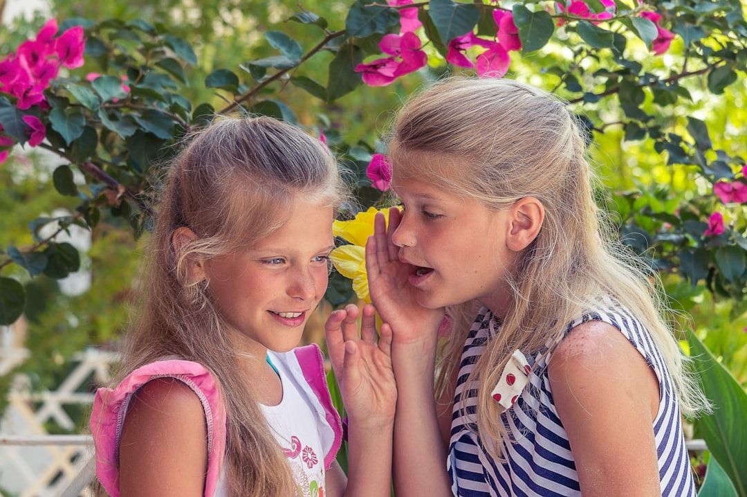 Zwei junge Mädchen stehen zwischen leuchtenden Blumen in Aachen und flüstern einander etwas zu. Ein Mädchen hält eine gelbe Blume an das Ohr ihrer Freundin.