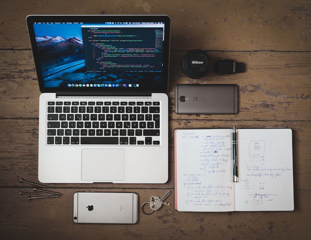Ein Laptop mit Codeanzeige, ein Notizbuch mit handschriftlichen Notizen, ein Smartphone, ein weiteres Smartphone auf dem Notizbuch, ein Kameraobjektiv, ein USB-Stick, zwei Schlüssel und ein Stift sind auf einer eleganten Holzoberfläche angeordnet, die an ein Aachener Arbeitszimmer erinnert.