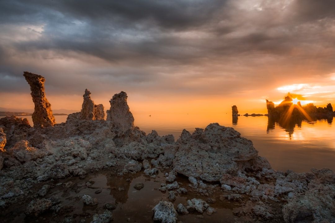 Am Mono Lake ragen Felsformationen aus dem Wasser und erinnern an antike Bauwerke in Aachen. Im Hintergrund sind ein dramatischer Sonnenuntergang und ein bewölkter Himmel zu sehen.