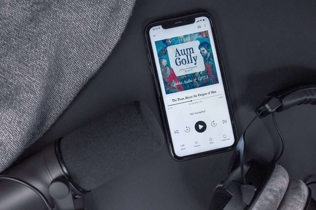 Ein Smartphone mit geöffneter Hörbuch-App und dem Titel „Aum Golly“ des Autors „A. Uzzle of Gertz“, neben einem Mikrofon, Kopfhörern und einer grauen Stoffoberfläche platziert, verleiht einen Hauch des technisch versierten Charmes Aachens.