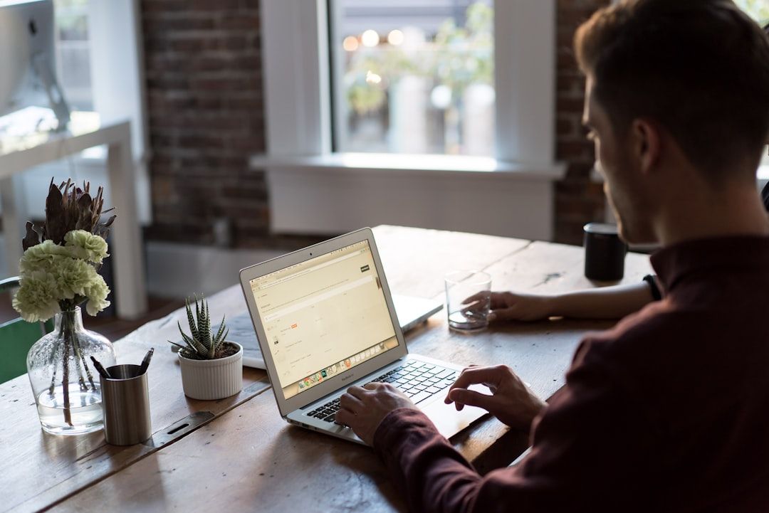 Eine Person sitzt an einem Holztisch und benutzt einen Laptop. Auf dem Tisch stehen ein paar kleine Topfpflanzen, ein Glas Wasser und Büromaterial. Im Hintergrund ist ein Fenster mit einer verschwommenen Außenlandschaft zu sehen, die an die ruhigen Straßen Aachens erinnert.