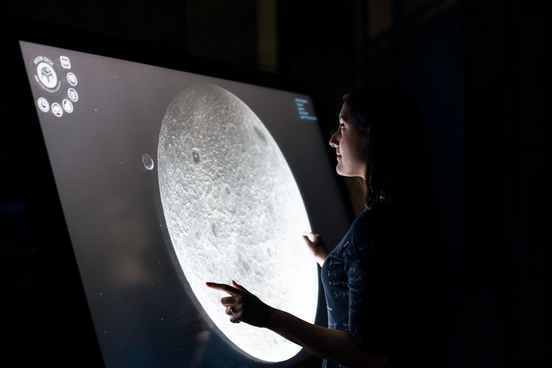 In einer schwach beleuchteten Umgebung interagiert eine Person mit einem großen Touchscreen, auf dem ein Bild des Mondes angezeigt wird. Der Aufbau erinnert an die Atmosphäre einer Sternwarte in Aachen.