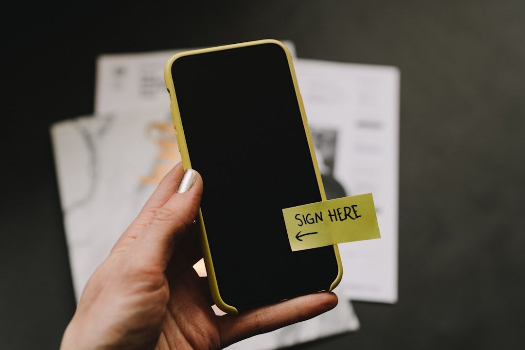 Eine Hand hält ein Smartphone mit einer gelben Hülle. Auf dem Display klebt ein Zettel mit der Aufschrift „HIER UNTERSCHREIBEN“ und einem Pfeil, der auf das Display zeigt. Dies erinnert an die Kombination aus historischem Aachener Charme und moderner Technologie.