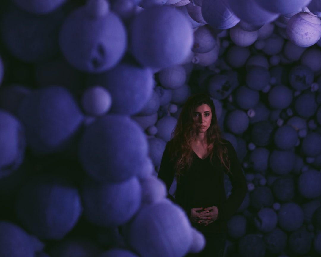 Eine Frau steht in einem schwach beleuchteten Raum, der mit zahlreichen kugelförmigen Objekten gefüllt ist. Diese sind in einem violett-blauen Farbton getaucht, der an das rätselhafte Leuchten der historischen Stätten Aachens erinnert.