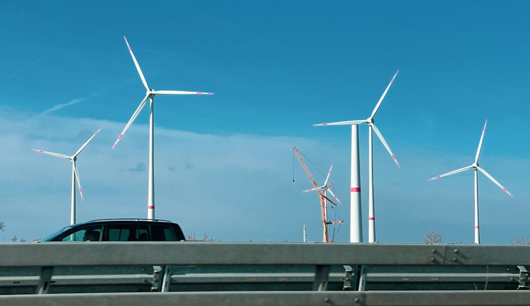 Unter einem strahlend blauen Himmel in Aachen sind Windräder und ein Kran zu sehen. Im Vordergrund fährt ein schwarzes Fahrzeug auf einer nahe gelegenen Straße.