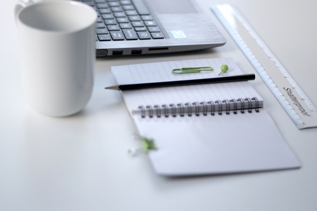 Eine weiße Kaffeetasse, ein Laptop, ein Notizblock mit Bleistift und grüner Büroklammer sowie ein Lineal liegen ordentlich angeordnet auf dem weißen Schreibtisch in meiner gemütlichen Aachener Wohnung.