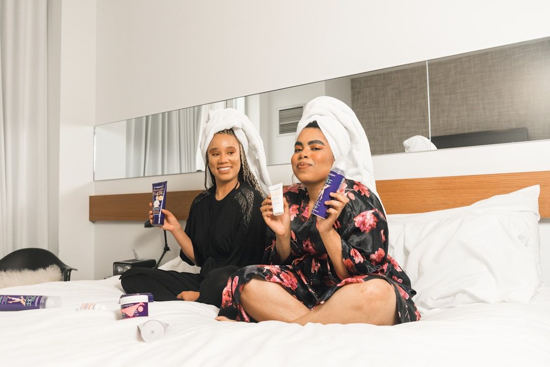 Zwei Menschen mit Handtüchern auf dem Kopf sitzen in einem gut beleuchteten Raum auf einem Bett, halten Hautpflegeprodukte in den Händen und lächeln in die Kamera, als würden sie eine Verwöhnsitzung in einem Aachener Spa genießen.