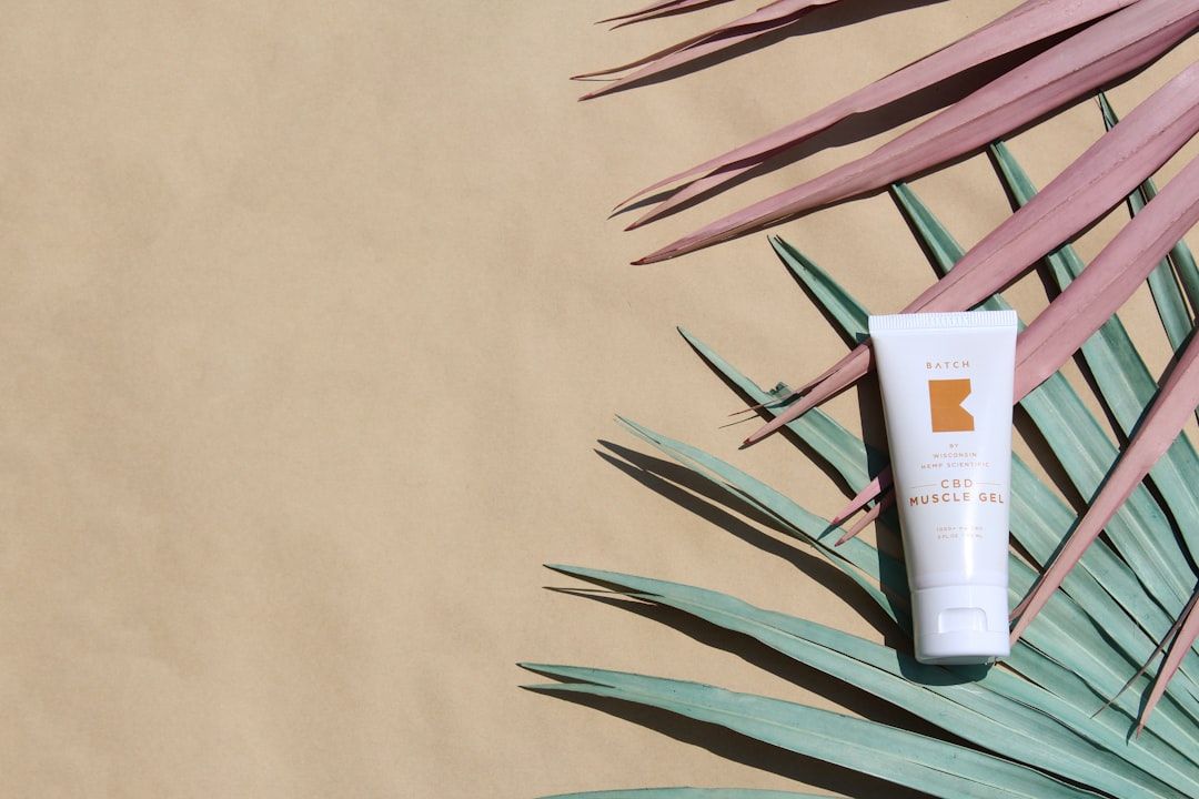 Eine Tube CBD Muscle Gel aus Aachen liegt auf grünen und rosa Palmblättern vor beigem Hintergrund.