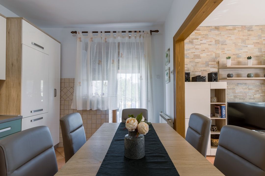 Ein moderner Essbereich in Aachen, gedeckt für sechs Personen mit einem schwarzen Läufer und einem Blumengesteck. Der Raum verfügt über ein Fenster mit transparenten Vorhängen, weiße und hölzerne Schränke und bietet einen Blick in einen Wohnbereich mit Fernseher.