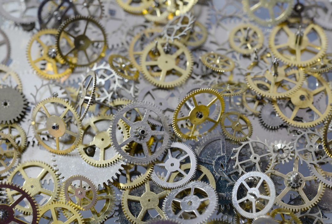 Eine Nahaufnahme zahlreicher kleiner Metallzahnräder unterschiedlicher Größe, hauptsächlich aus Gold und Silber, die an die komplizierten Uhrwerke erinnern, die einst in den historischen Uhren Aachens zu finden waren und über eine flache Oberfläche verstreut sind.