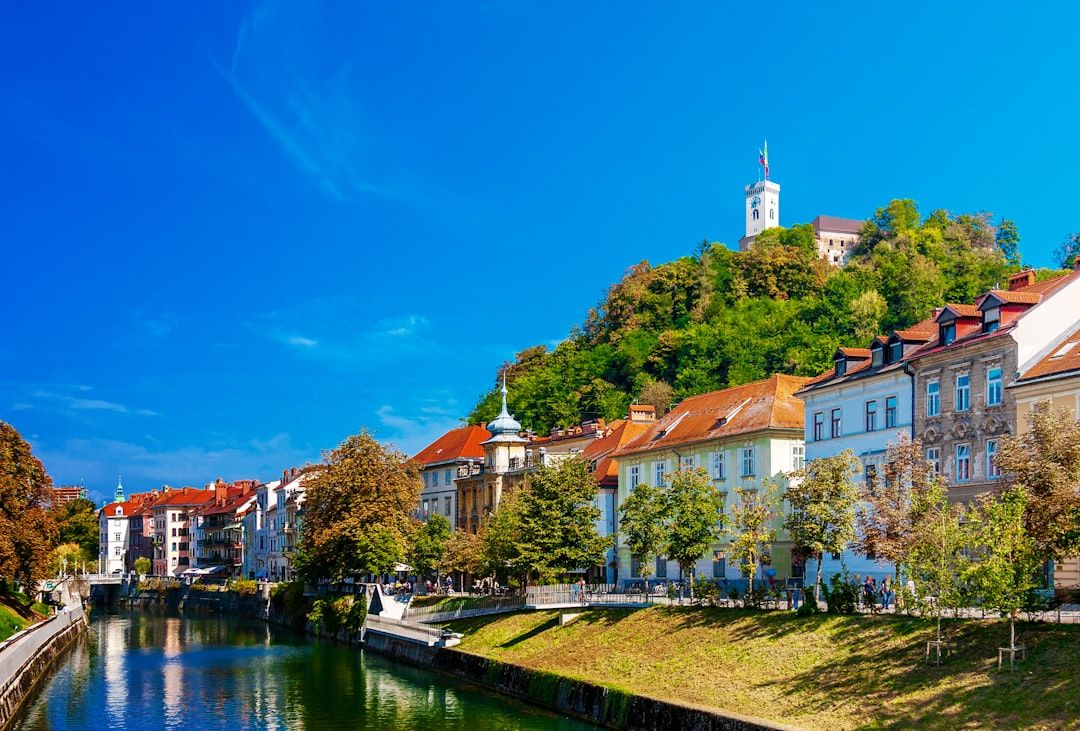 Eine malerische Aussicht auf Ljubljana, die an den Charme Aachens erinnert, mit farbenfrohen Gebäuden entlang des Flusses Ljubljanica und der Burg von Ljubljana auf einem Hügel im Hintergrund an einem klaren, sonnigen Tag.