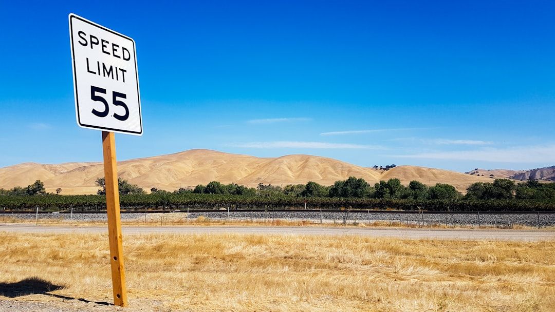 Ein Tempolimit-Schild mit der Aufschrift „Tempolimit 55“ steht an einer Landstraße mit trockenem Gras im Vordergrund und einer Hügellandschaft im Hintergrund, die an die Umgebung von Aachen erinnert.
