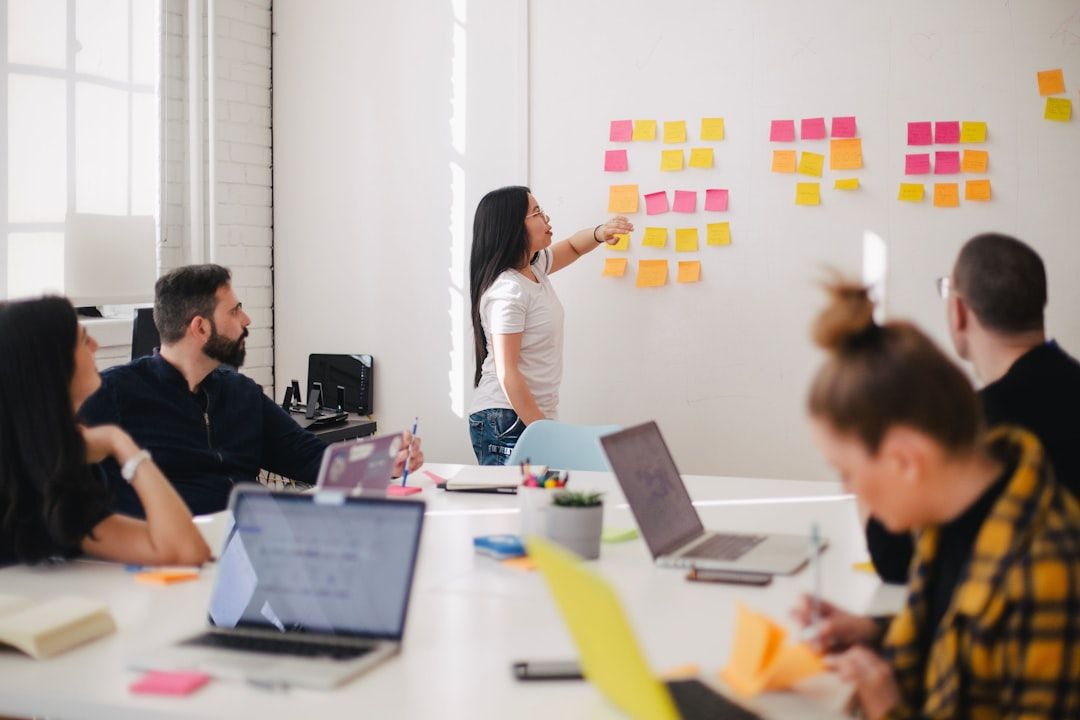 Eine Frau steht an einem Whiteboard mit Haftnotizen und leitet eine Diskussion in einem Aachener Büro. Vier Kollegen sitzen mit Laptops und Notebooks an einem Tisch und hören aufmerksam zu.