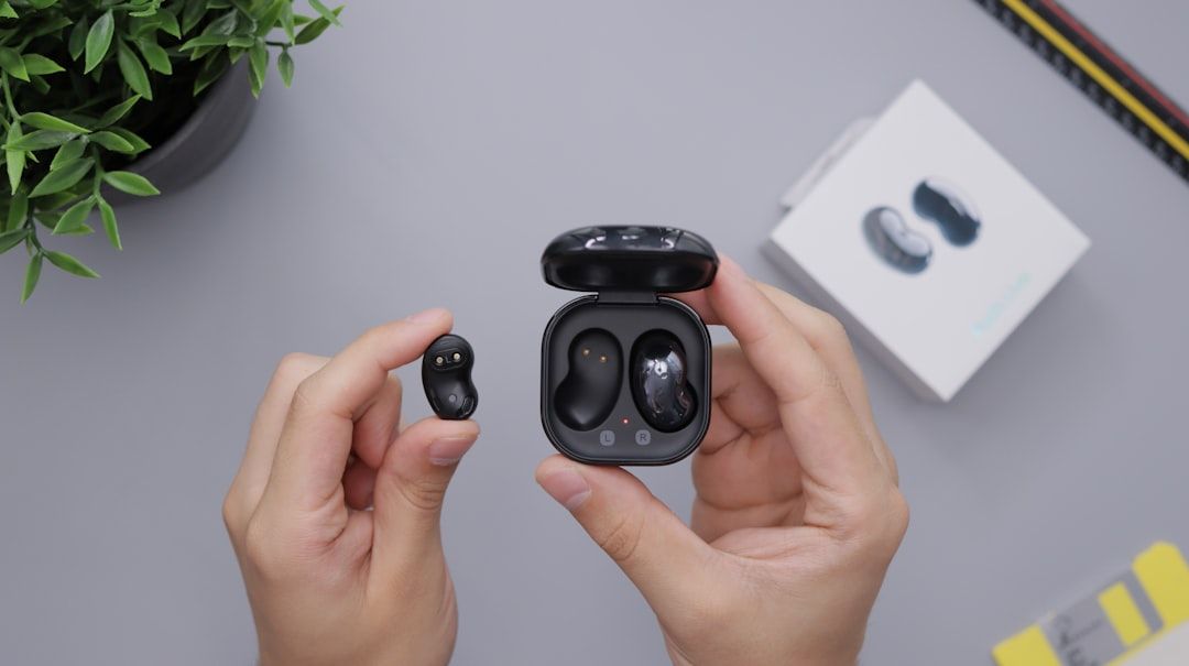 Hände halten schwarze kabellose Ohrhörer und ein Ladeetui, mit Produktverpackungen und einer Pflanze auf dem Tisch, was an ein elegantes Design erinnert, wie man es in Aachener Technologiezentren finden würde.