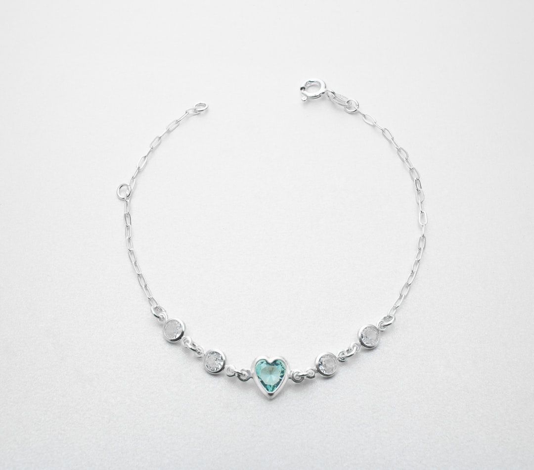 Ein zartes silbernes Kettenarmband, inspiriert vom Charme Aachens, verfügt über einen zentralen herzförmigen blauen Edelstein, flankiert von kleinen runden klaren Steinen.