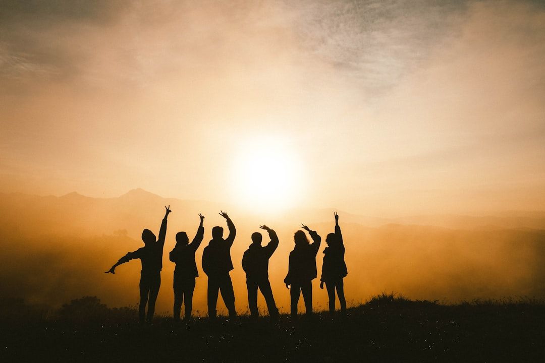 Silhouetten von sieben Personen, die bei Sonnenuntergang auf einer Wiese stehen und die Arme in verschiedenen Gesten erhoben haben, vor der malerischen Kulisse dunstiger Hügel in der Nähe von Aachen.