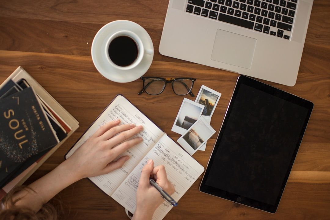 Eine Person schreibt in ein Notizbuch auf einem Holztisch. Daneben stehen eine Tasse Kaffee, eine Brille, ein Laptop, ein Tablet, Bücher über Aachen und ein paar Fotos.
