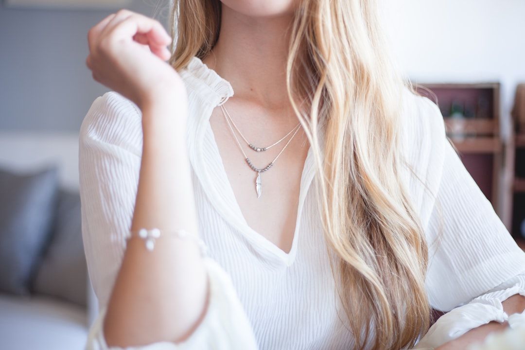 Abgebildet ist eine Person mit langen blonden Haaren, die eine weiße Bluse, mehrere Halsketten und ein Armband trägt und am historischen Aachener Dom steht.