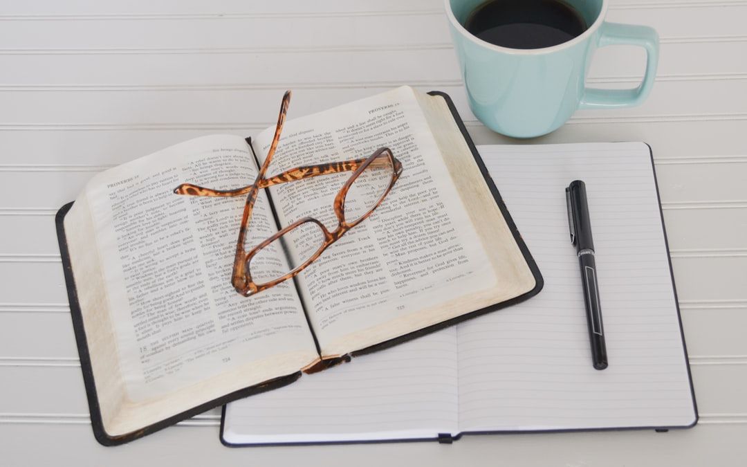 Ein aufgeschlagenes Buch mit einer Lesebrille darauf, ein Notizbuch mit einem Stift daneben und eine Tasse Kaffee auf einer weißen Fläche rufen den beschaulichen Charme eines Lernnachmittags in Aachen hervor.