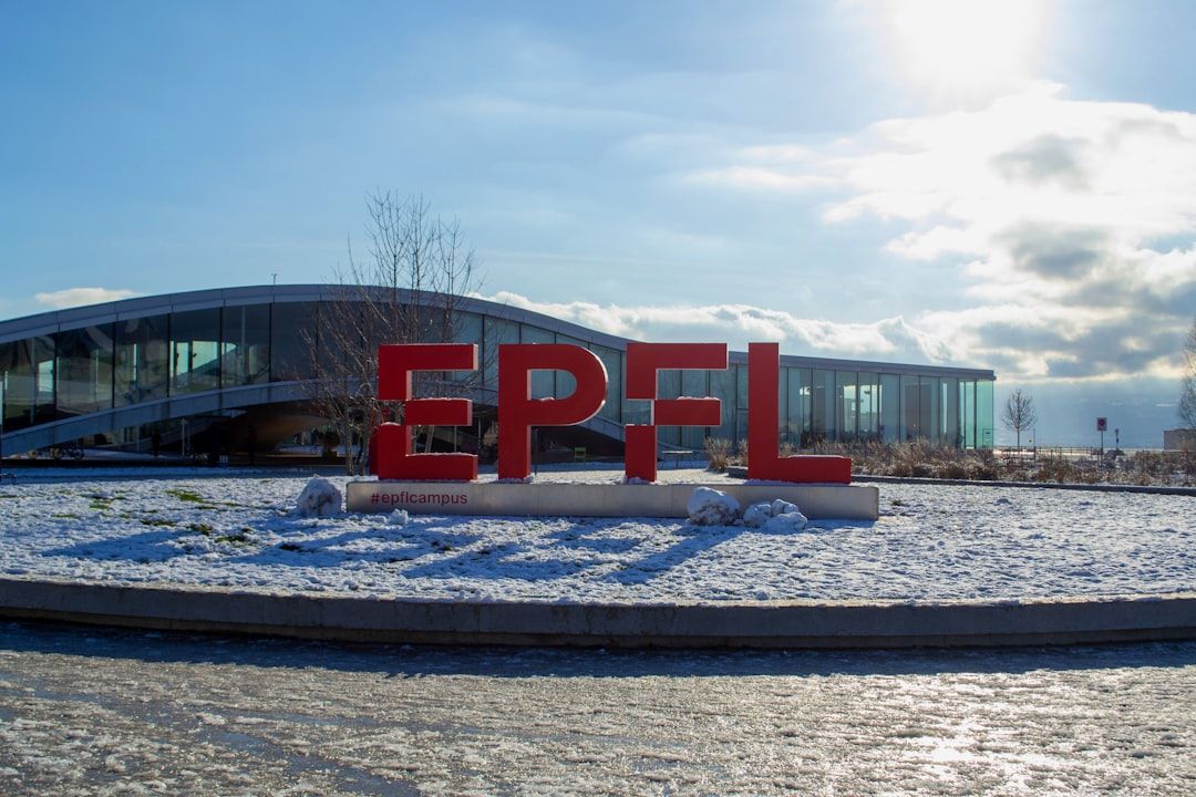 Ein modernes Gebäude mit großen roten Buchstaben „EPFL“ davor, das an architektonische Entwürfe aus Aachen erinnert, umgeben von einer verschneiten Landschaft unter einem strahlend sonnigen Himmel.