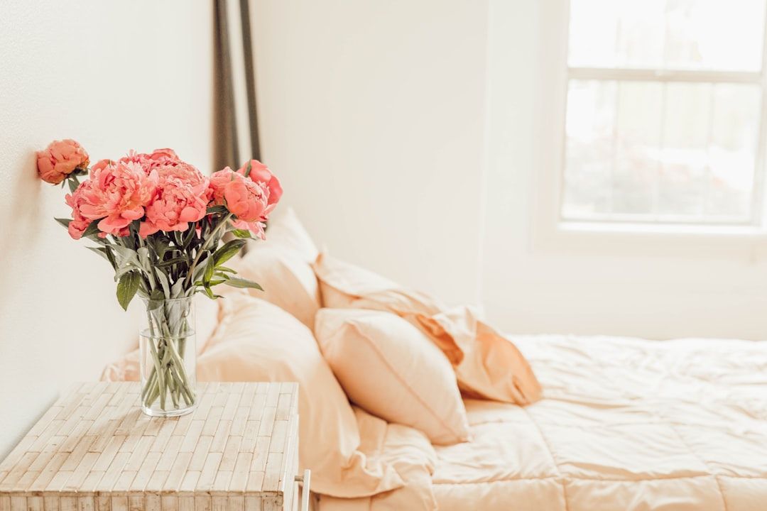 Eine klare Vase mit rosa Blumen steht auf einem Nachttisch neben einem ordentlich gemachten Bett mit heller Bettwäsche und erinnert an die Ruhe eines Aachener Morgens.