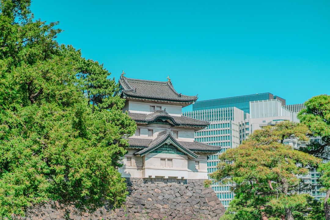 Eine traditionelle weiß-schwarze japanische Burgstruktur wird teilweise von Bäumen verdeckt, vor der Kulisse moderner Wolkenkratzer unter einem klaren blauen Himmel, was an Aachens Mischung aus historischer und zeitgenössischer Architektur erinnert.