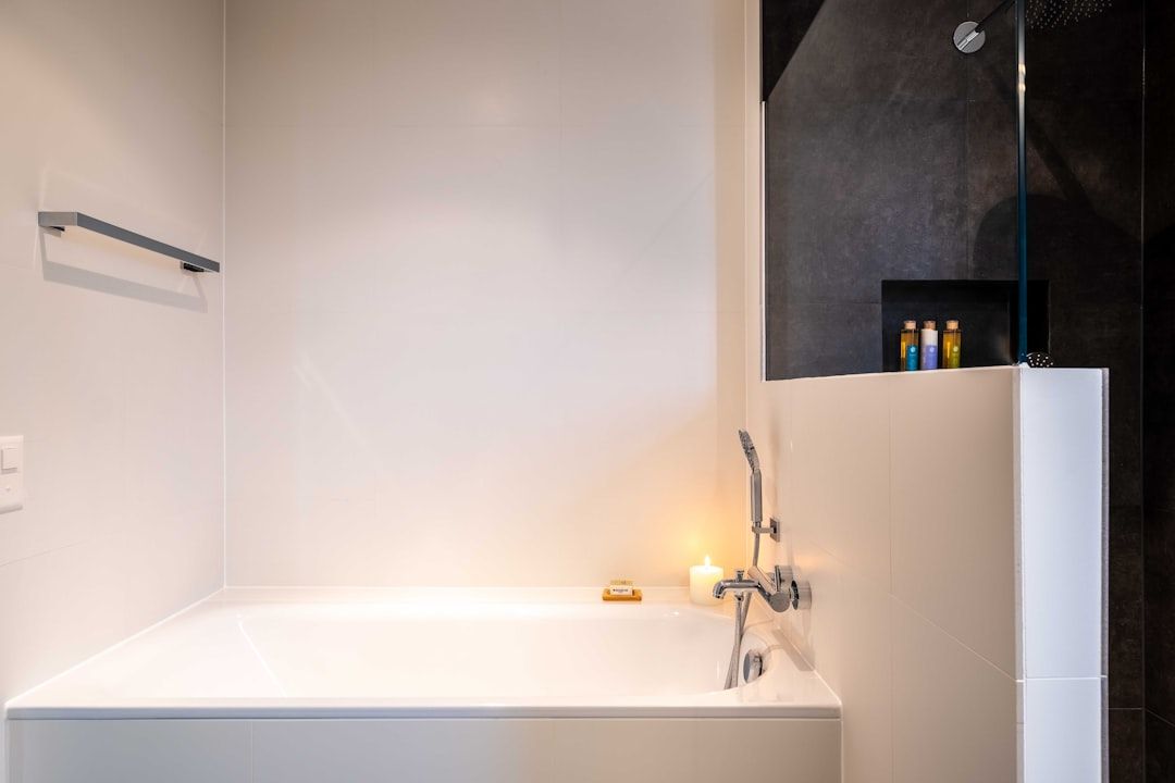 Minimalistisches Badezimmer mit weißer Badewanne, brennender Kerze, Seifenschale und modernem Duschbereich mit schwarzen Fliesen, die an das elegante Design Aachens erinnern, komplett mit verschiedenen Toilettenartikeln auf einem kleinen Regal.