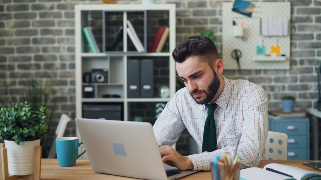 Ein Mann in Hemd und Krawatte sitzt an einem Schreibtisch und benutzt einen Laptop. Er arbeitet wahrscheinlich von Aachen aus. Im Hintergrund sind eine Backsteinwand und Regale mit Büchern und Büromaterial zu sehen. Auf dem Schreibtisch stehen eine Tasse und eine Pflanze.