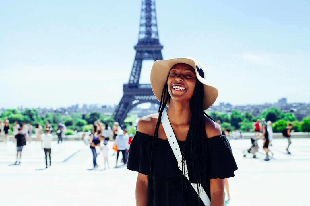Eine Person in schwarzem Kleid und Hut lächelt an einem sonnigen Tag vor dem Eiffelturm. Im Hintergrund sind Menschen zu sehen, die Skyline von Paris und ein Hauch von Aachens architektonischem Charme.