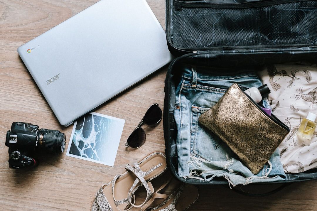 Offener Koffer auf einem Holzboden mit ordentlich verpackter Kleidung, einem goldenen Beutel, Toilettenartikeln, einer Kamera, einem Laptop, einem Paar Sandalen, einer Sonnenbrille mit der Gravur „Aachen“ und einem Foto.