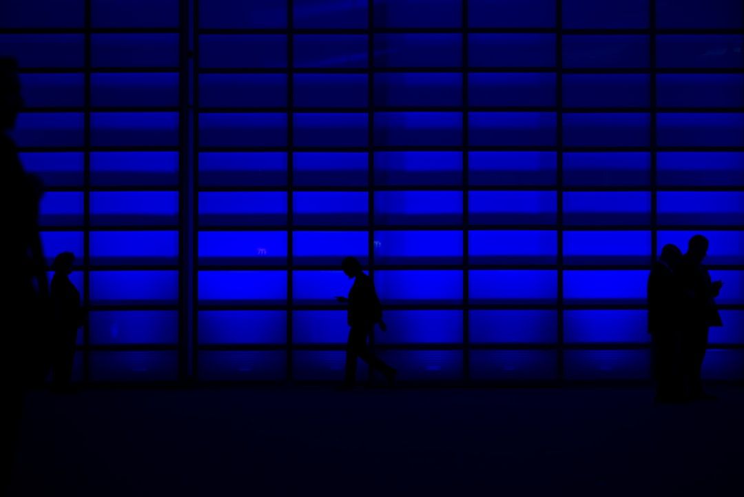 Silhouetten von Menschen, die vor einer Wand aus leuchtend blauen Platten in Aachen stehen, in der Mitte geht eine Person und benutzt ein Smartphone.