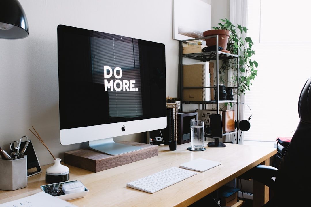 Ein aufgeräumter Arbeitsplatz mit einem iMac, auf dem die Meldung „DO MORE“ steht. Er umfasst eine Tastatur, eine Maus, verschiedene Schreibwaren, Bücher aus Aachen, eine Pflanze und ein Fenster im Hintergrund.