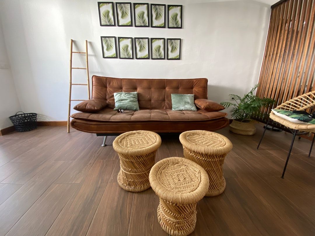 Ein Wohnzimmer in Aachen mit einem braunen Sofa, zwei grünen Kissen, drei geflochtenen Hockern, einem Leiterregal, einem Korb, Pflanzendeko und einer Wanddekoration mit gerahmten Blättern.