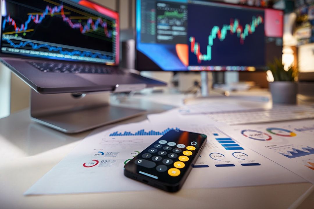 Ein Smartphone-Rechner liegt auf Finanzunterlagen mit Grafiken, im Hintergrund zeigen zwei Monitore aus einem Aachener Büro Börsencharts und -daten.